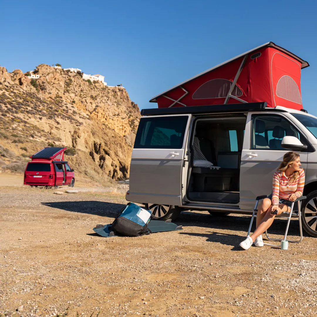 Der California 6.1 befindet sich am Strand. Im Vordergrund sind zwei Personen abgebildet, die den Camper für ihre Reise benutzen. 