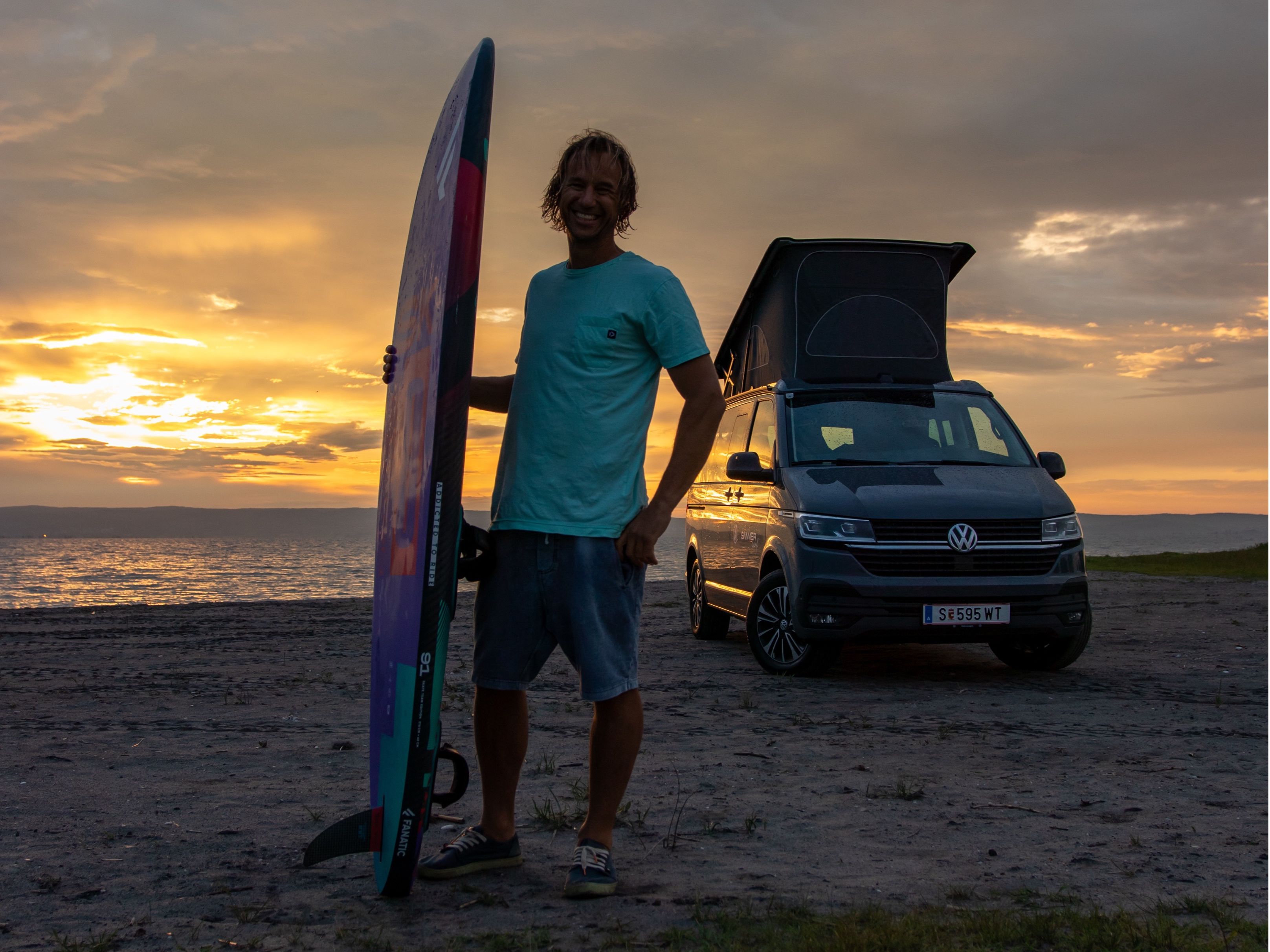 Chris Sammer steht bei Sonnenuntergang mit seinem Surfboard am Strand, sein VW California parkt mit ausgeklapptem Dach dahinter