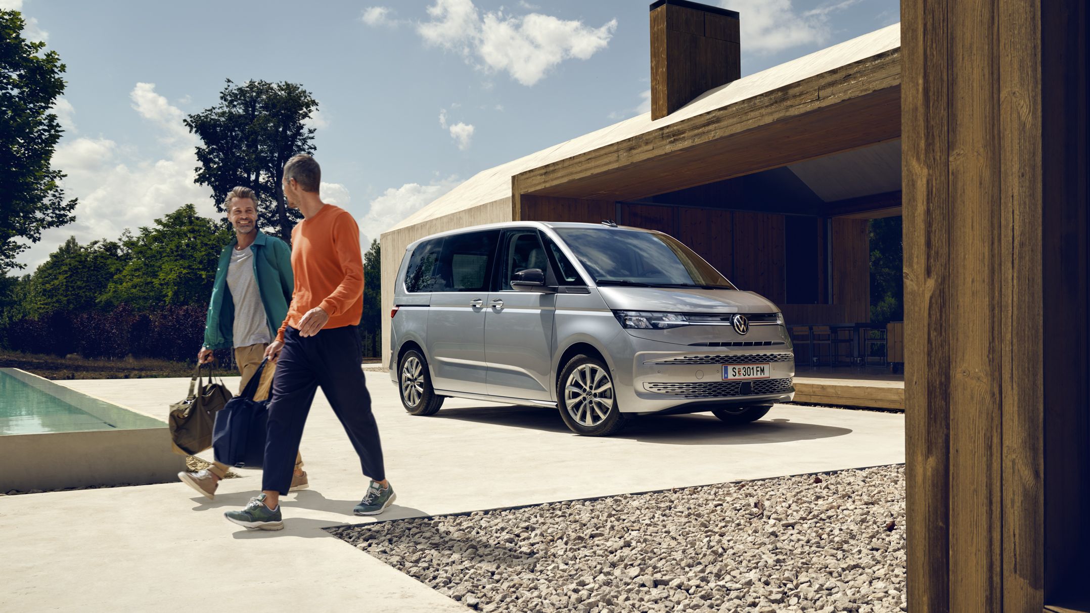 Ein Volkswagen Multivan in Silber parkt vor einem Haus mit Pool, zwei Männer mit Sporttaschen bewegen sich von dem Fahrzeug fort.