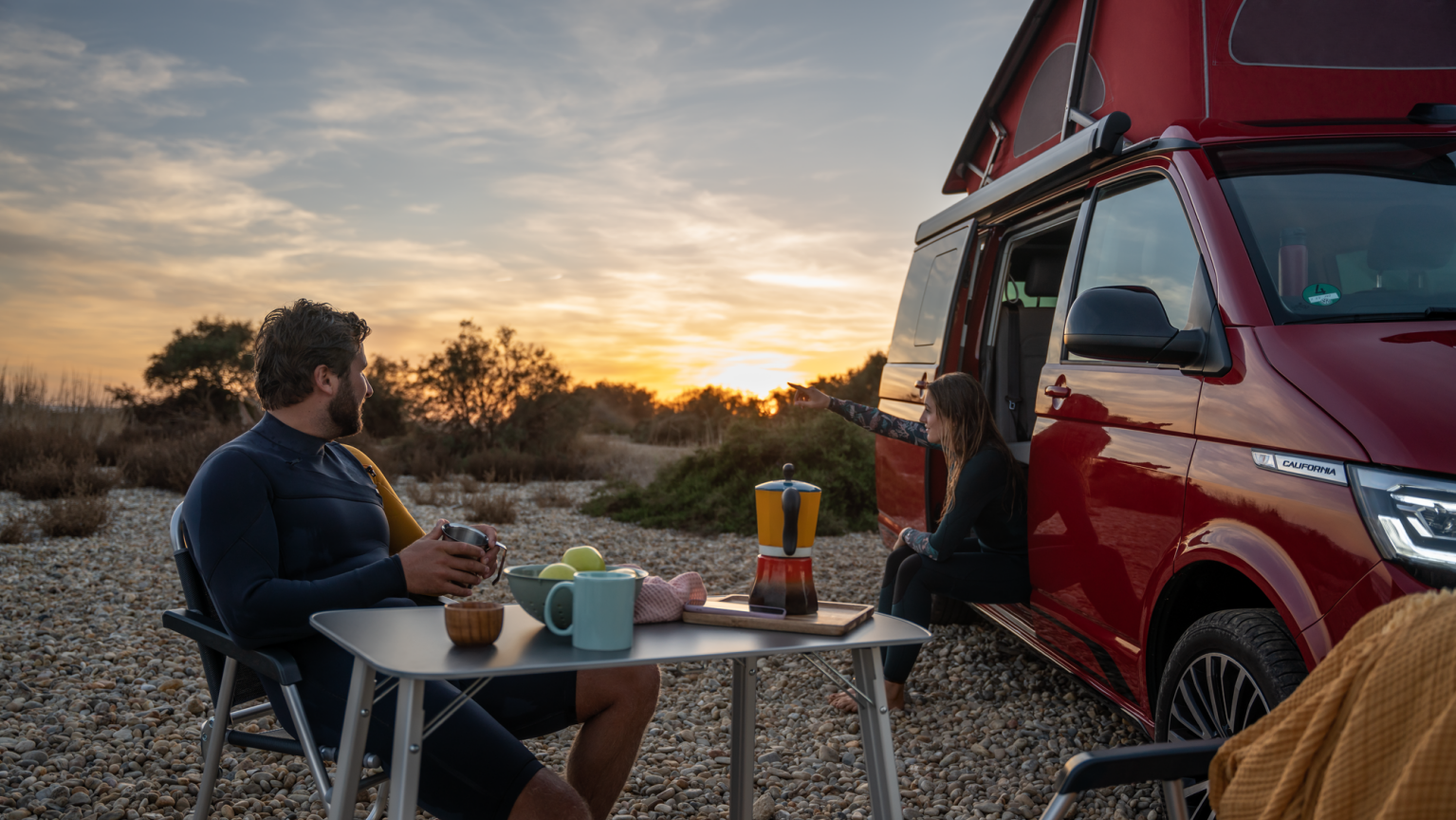 Ein Volkswagen California in tot, eine Frau sitzt an der Kante des Einstiegs und ein Mann an einem Camping Tisch davor