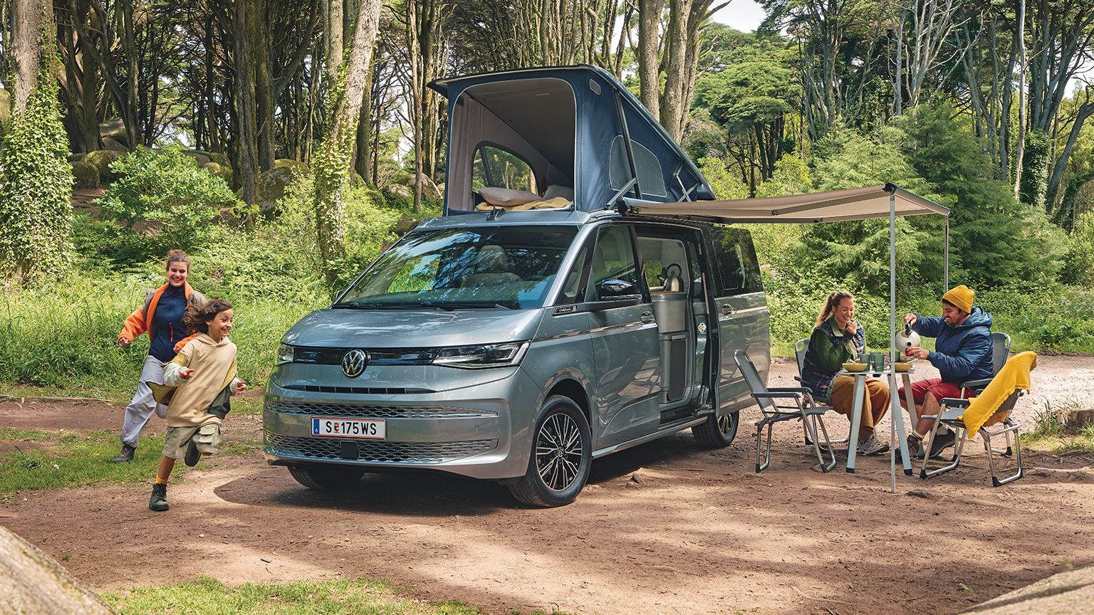 Ein New Multivan parkt im Wald,  zwei Kinder laufen darum herum, die Eltern frühstücken unter der ausgefahrenen Marquise