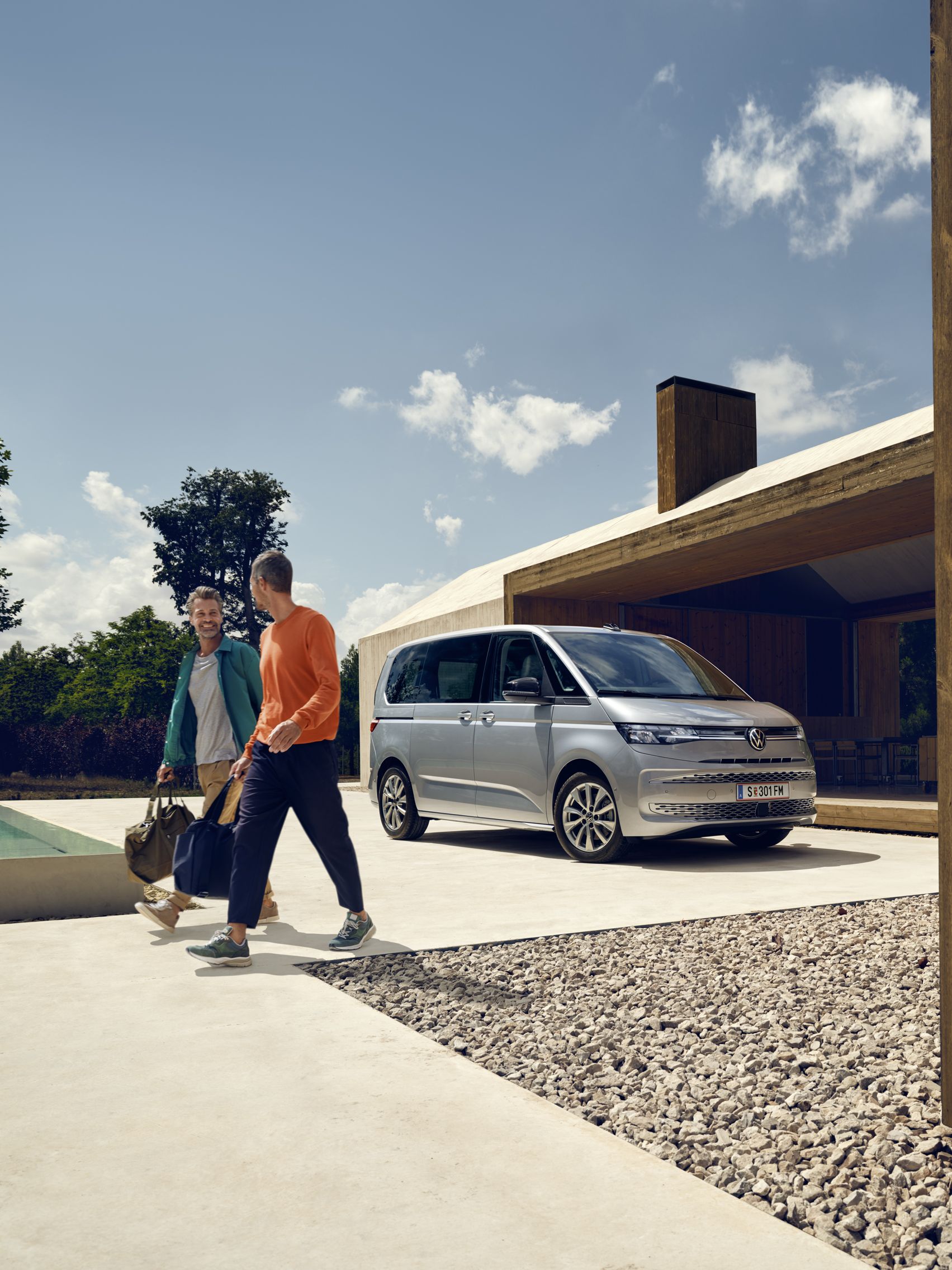 Ein Volkswagen Multivan in Silber parkt vor einem Haus mit Pool, zwei Männer mit Sporttaschen bewegen sich von dem Fahrzeug fort.