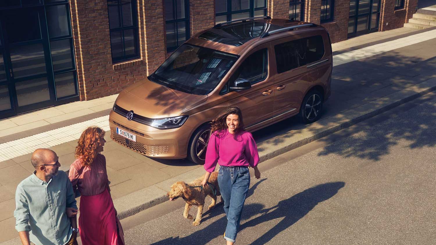 VW Caddy in copperbronze steht in der Stadt. Vater und Tochter gehen vom Fahrzeug weg.