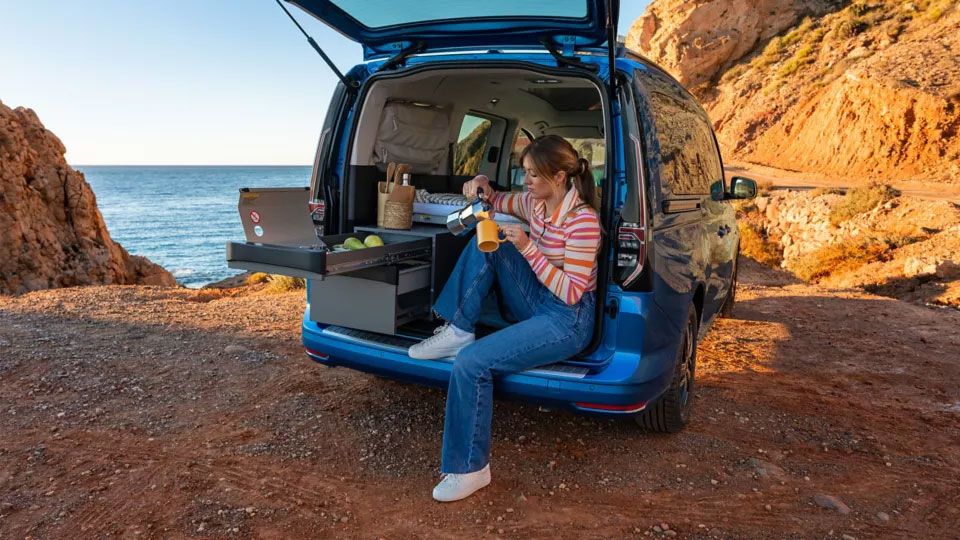 Eine Frau sitzt bei geöffneter Heckklappe in ihrem VW Caddy California, der am Meer parkt