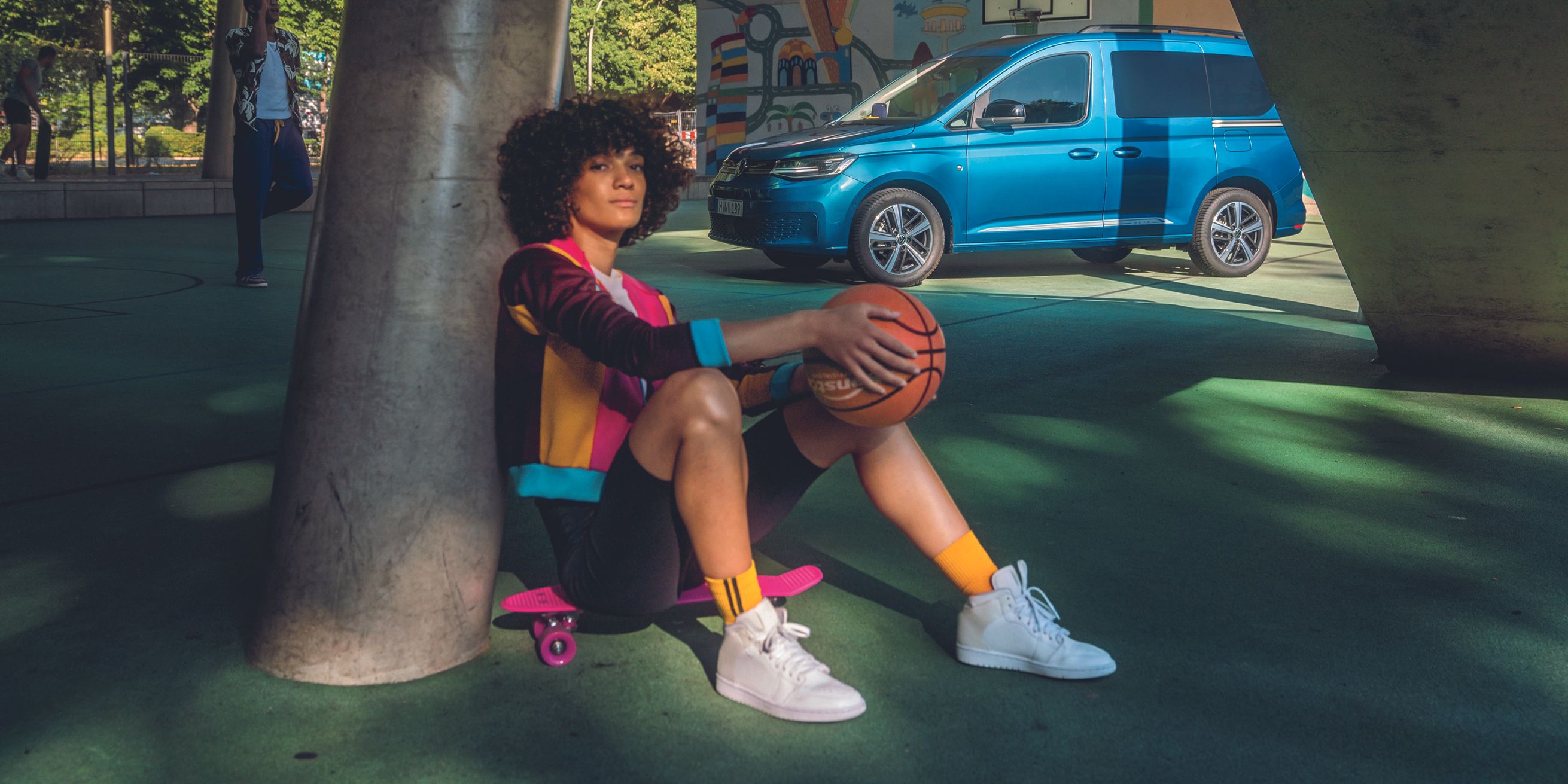 Ein blauer VW Caddy parkt auf einem Basketballplatz, eine Spielerin sitzt auf einem Skateboard davor