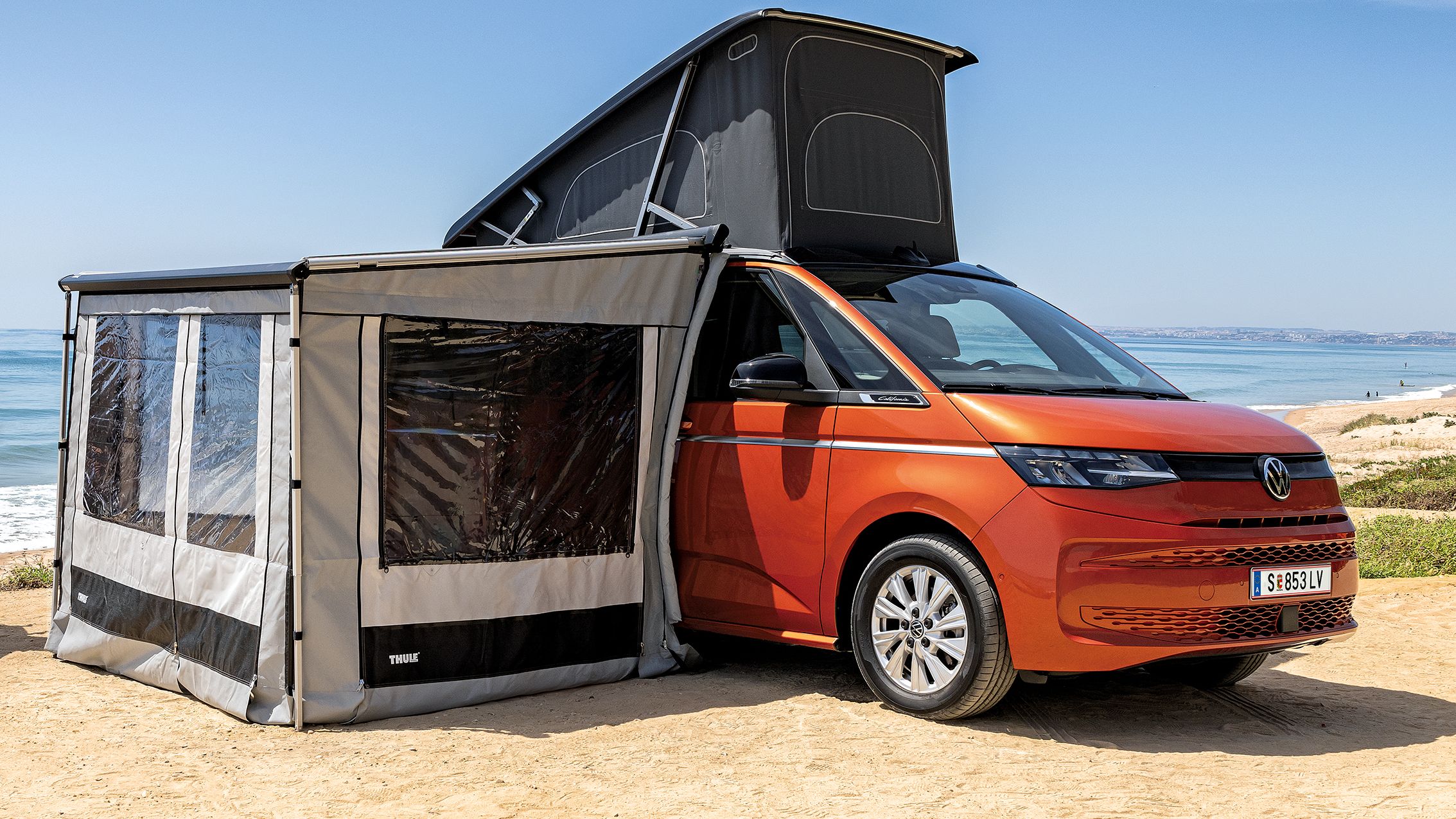 Zwei Surfer laufen an ihrem New California mit ausgeklapptem Dach und angebautem Zelt am Strand vorbei