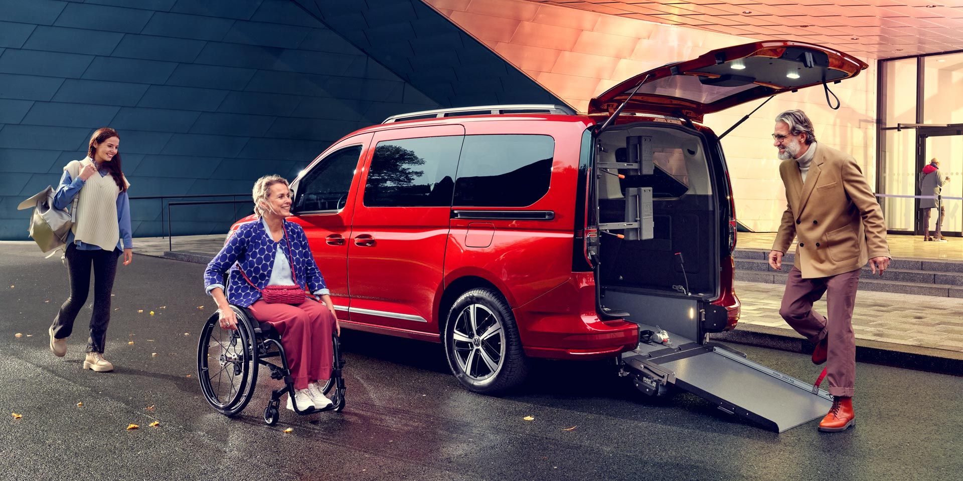 Ein roter VW Caddy mit offener Heckklappe und Rampe, eine Frau im Rollstuhl fährt darauf zu