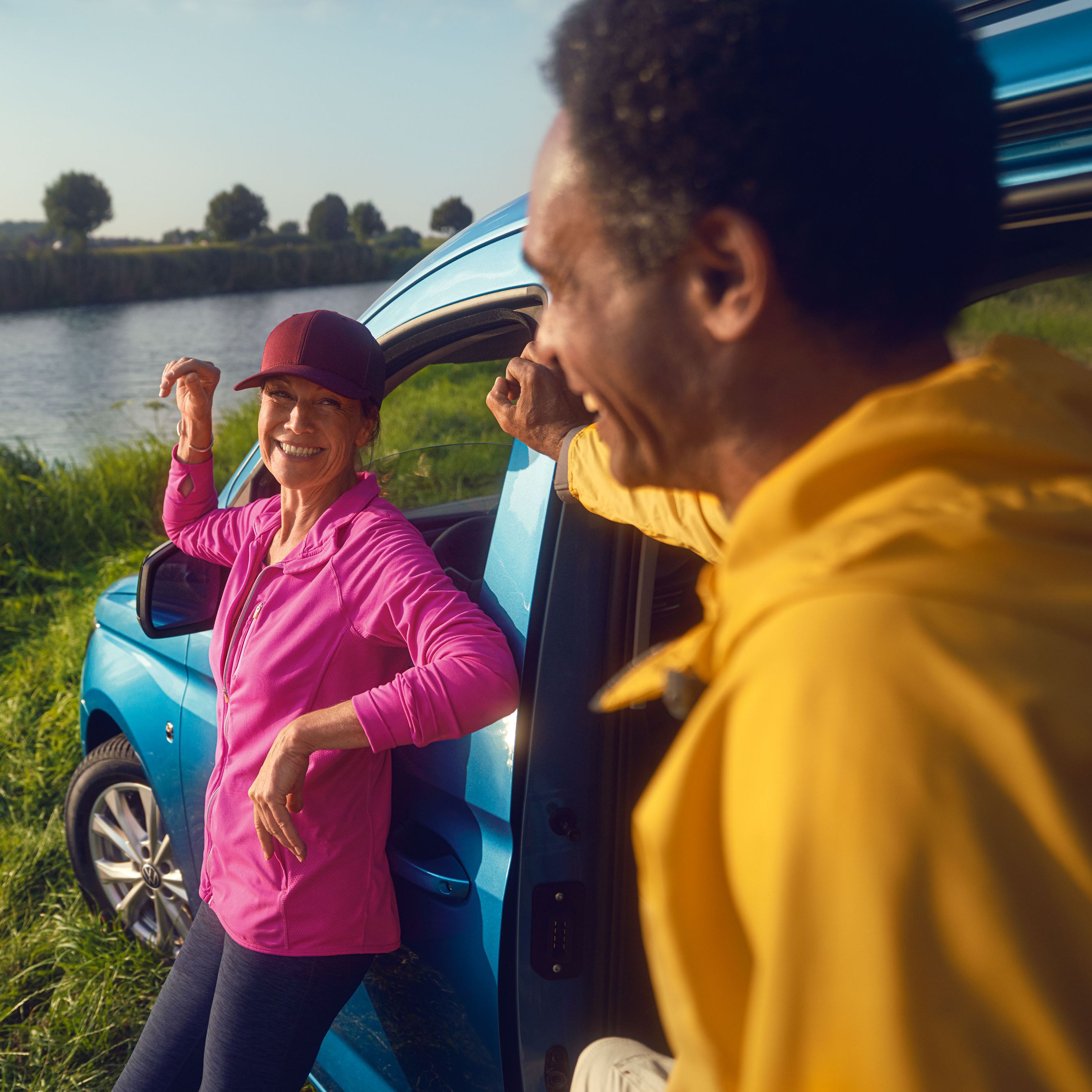 Mann und Frau lehnen an Seite eines blauen VW Caddys, der an einem See steht