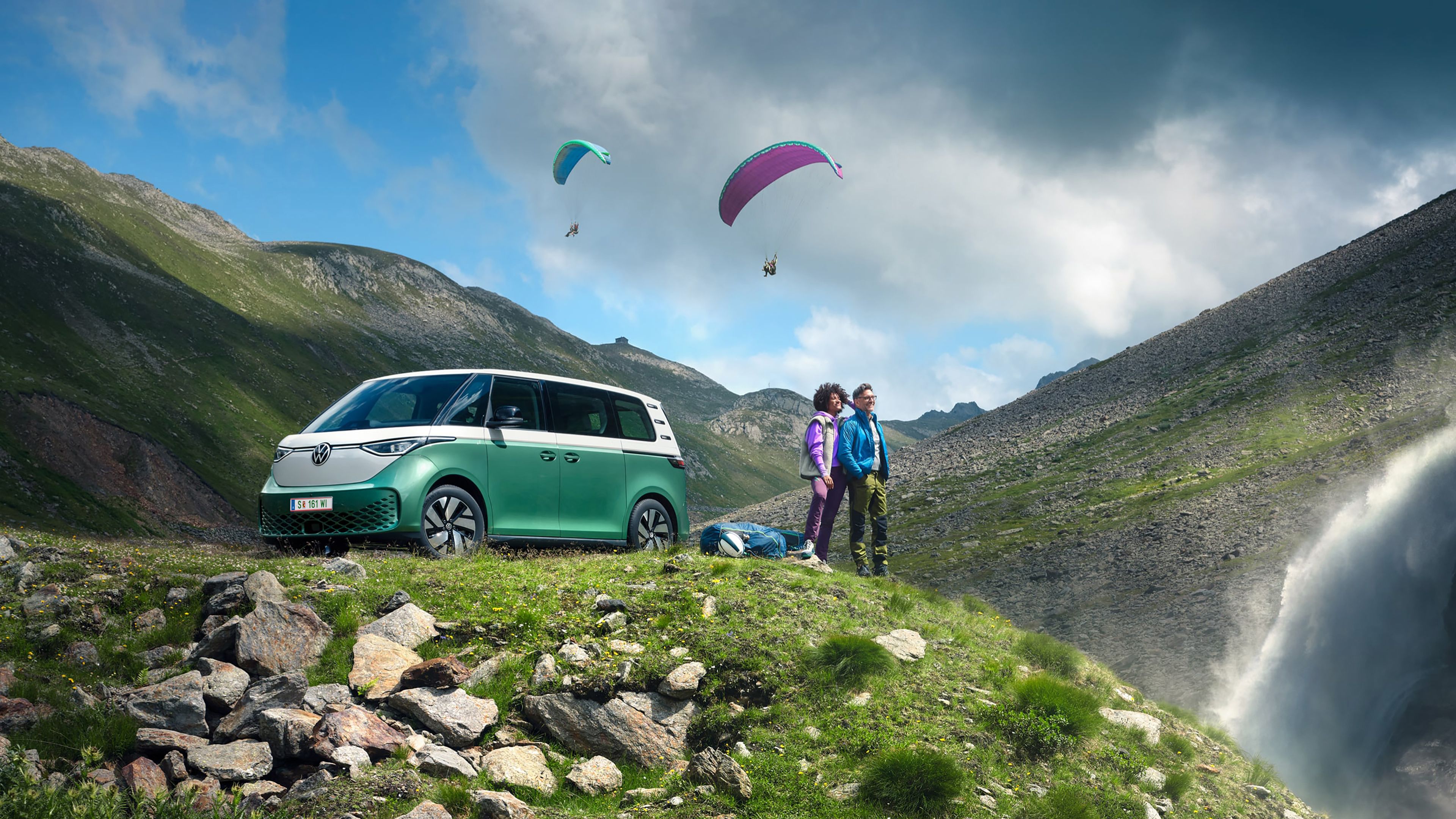 Ein grüner Volkswagen ID. Buzz parkt an einer Bergstraße, ein Mann und eine Frau beobachten Paragleiter.