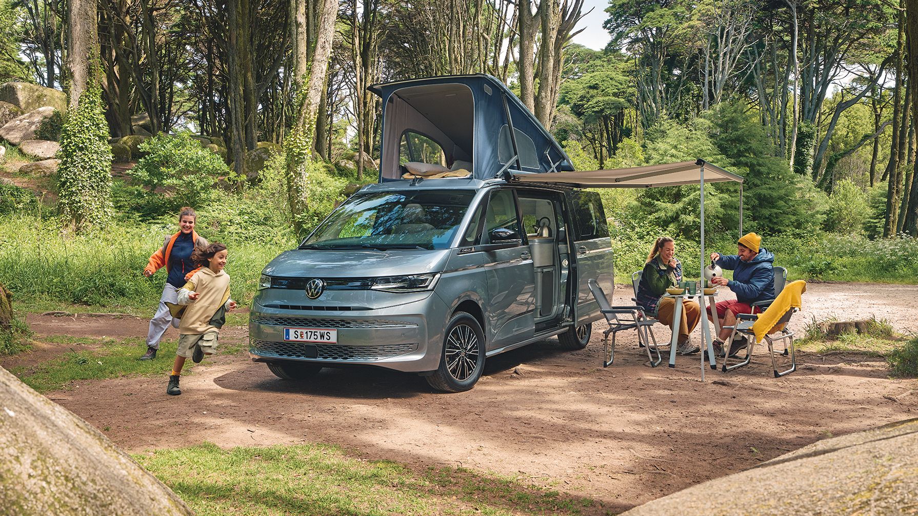 Ein New Multivan parkt im Wald,  zwei Kinder laufen darum herum, die Eltern frühstücken unter der ausgefahrenen Marquise