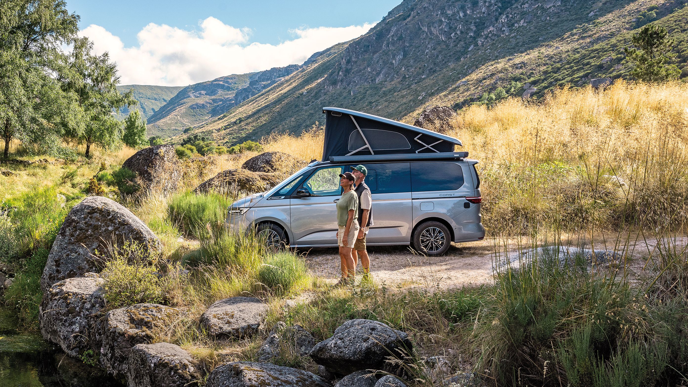 Ein New California mit ausgeklapptem Dach parkt an einem See, ein Paar steht am Seeufer