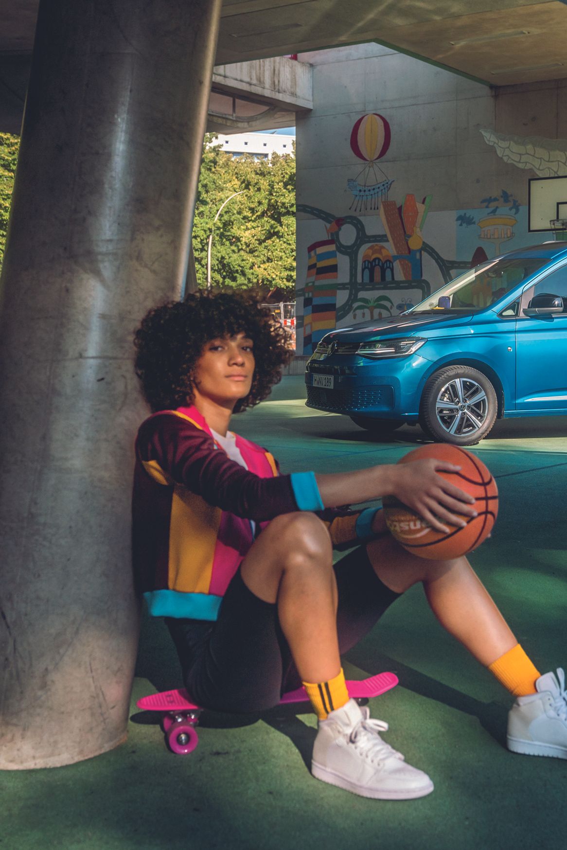 Ein blauer VW Caddy parkt auf einem Basketballplatz, eine Spielerin sitzt auf einem Skateboard davor