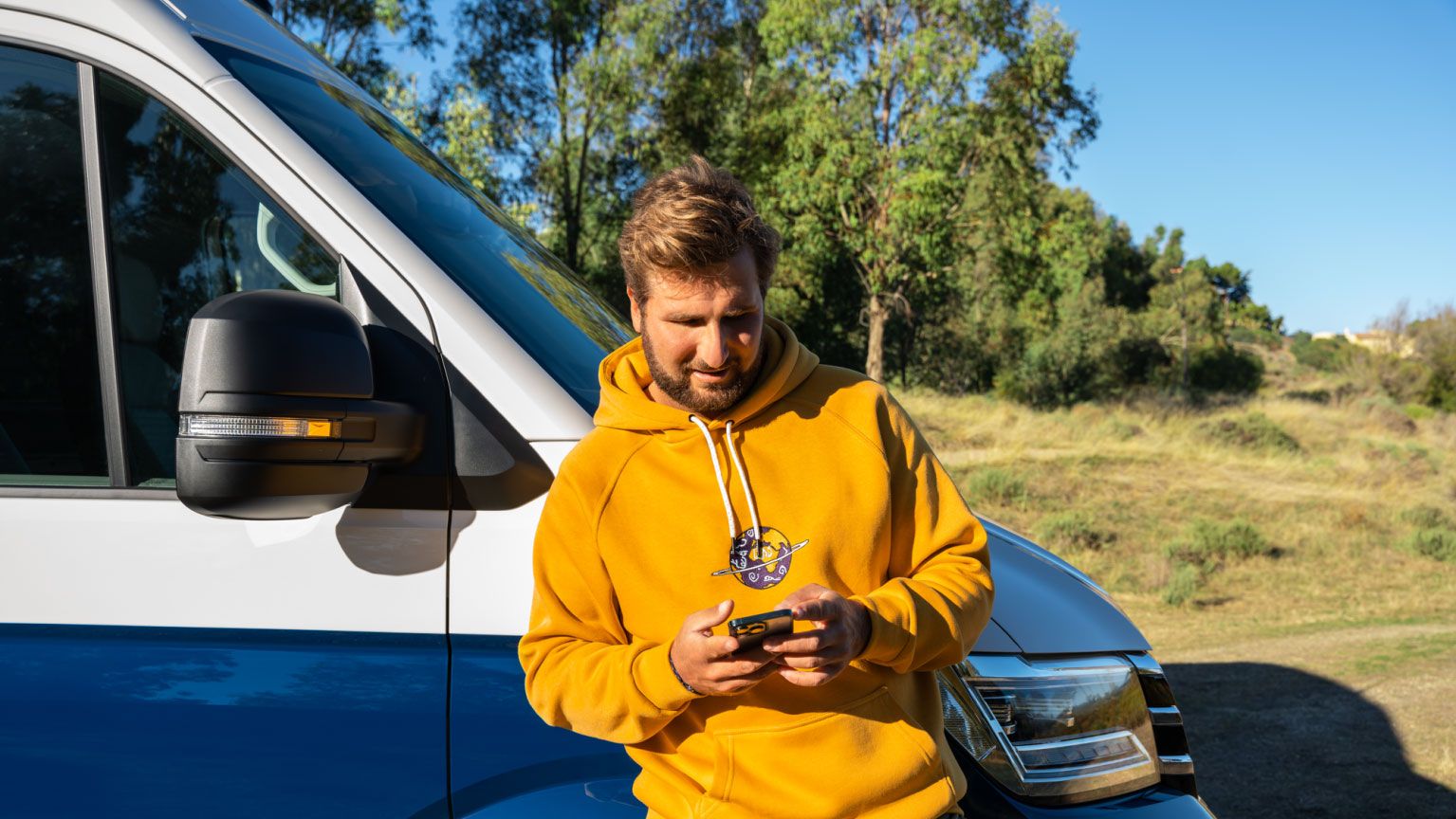 Ein Mann lehnt an seinem VW Nutzfahrzeug und blickt auf sein Smartphone