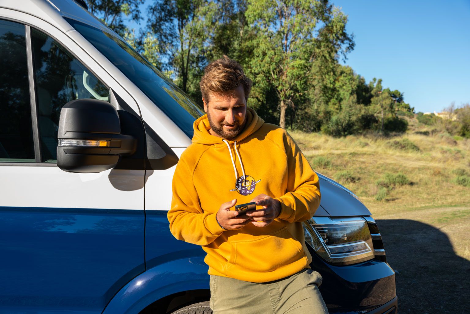 Ein Mann lehnt an seinem VW Nutzfahrzeug und blickt auf sein Smartphone