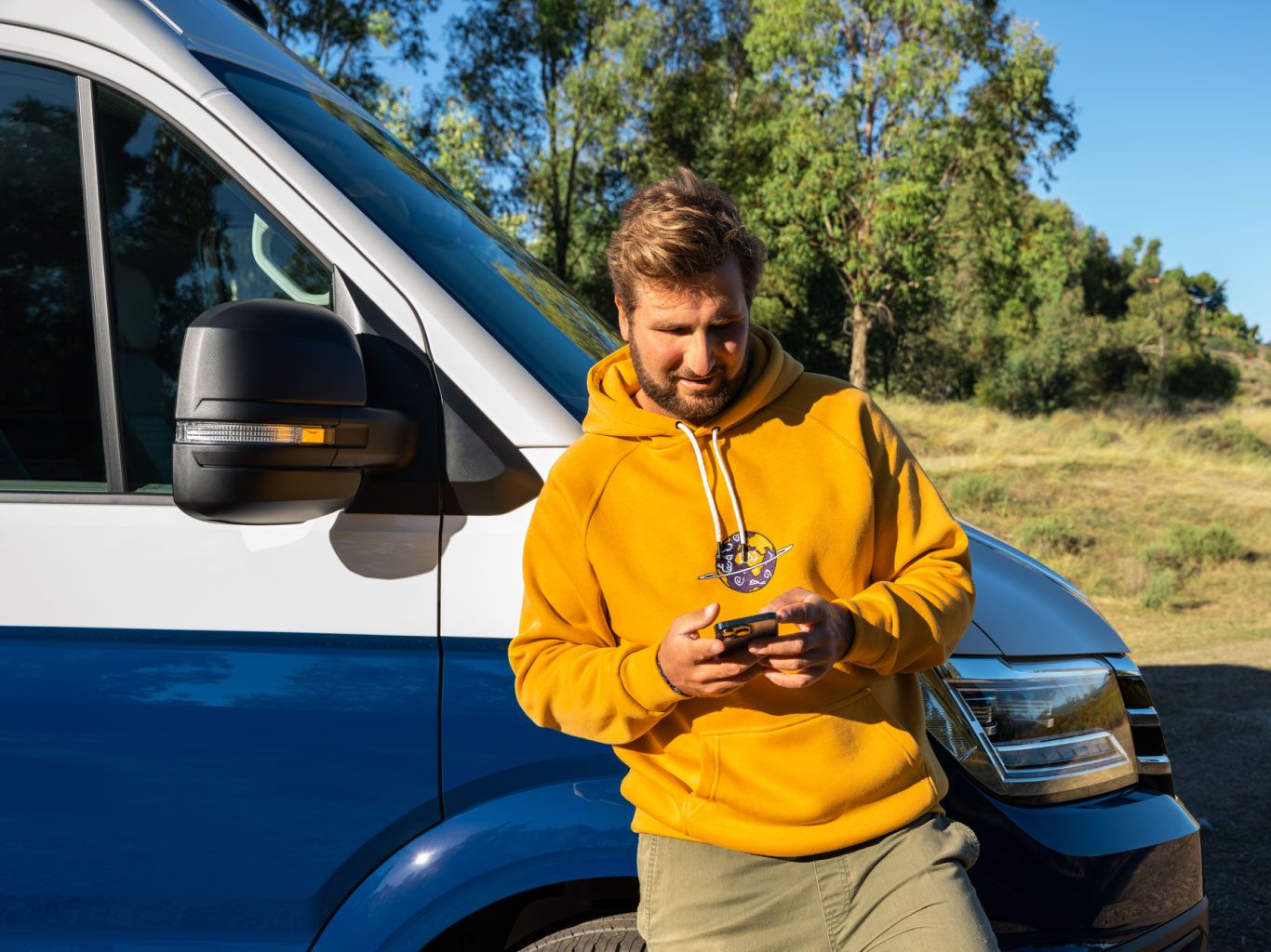 Ein Mann lehnt an seinem VW Nutzfahrzeug und blickt auf sein Smartphone