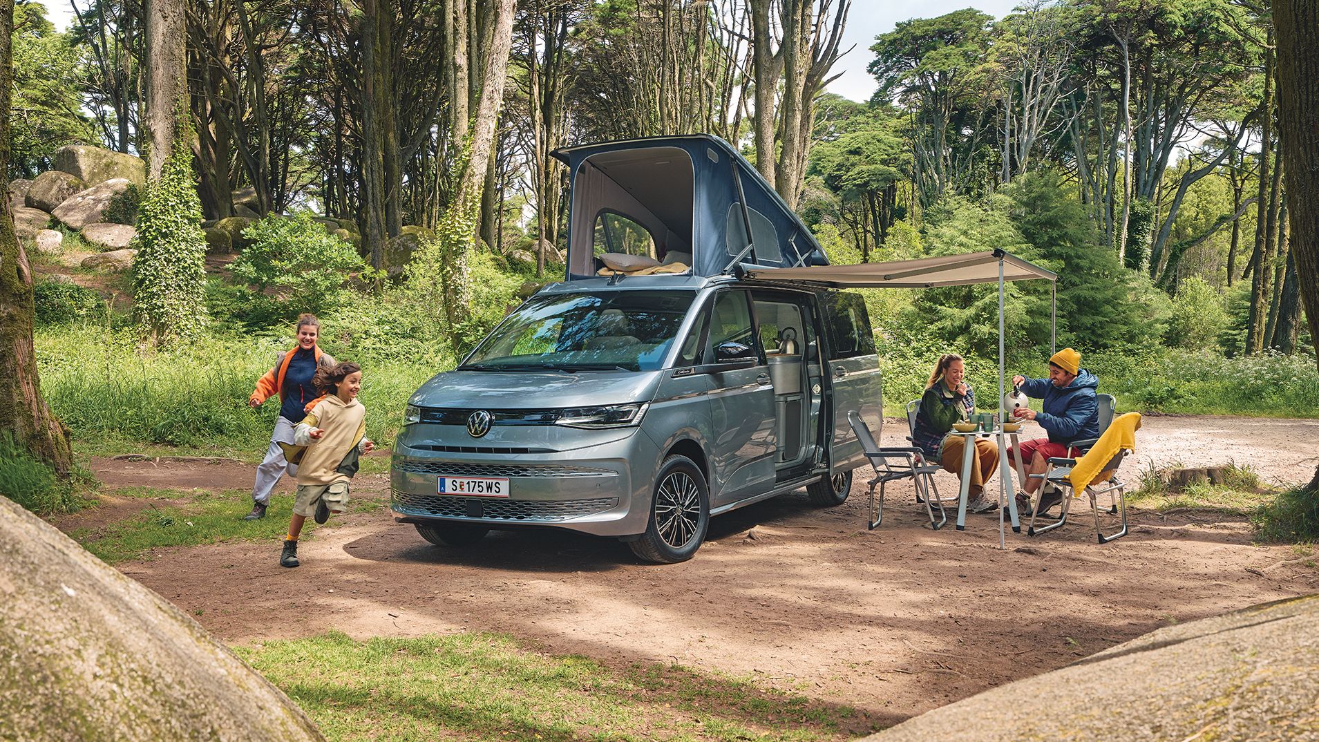 Ein New Multivan parkt im Wald,  zwei Kinder laufen darum herum, die Eltern frühstücken unter der ausgefahrenen Marquise