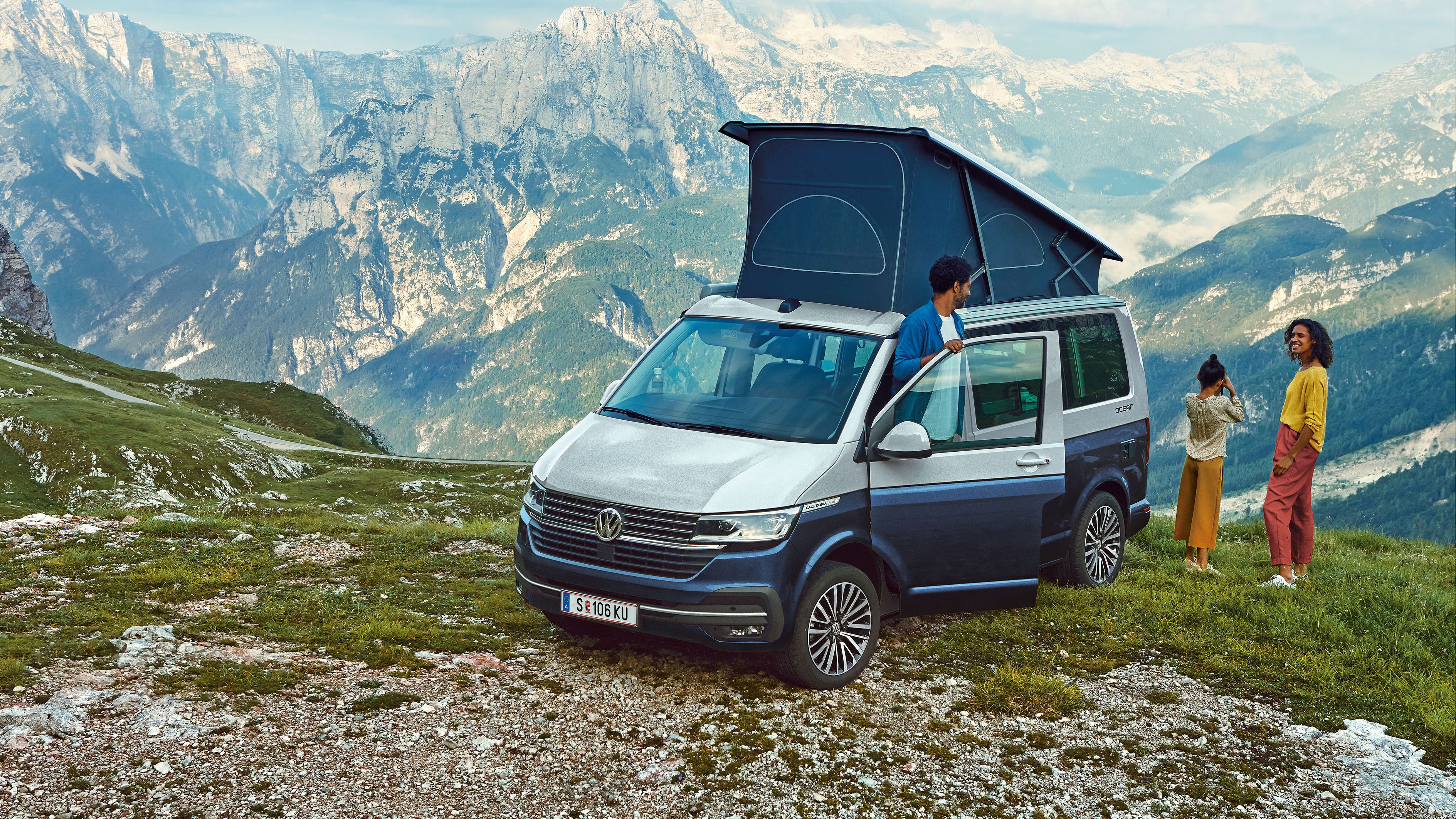 VW California 6.1 mit aufgestelltem Dach vor Bergpanorama, Familie steht nebenbei