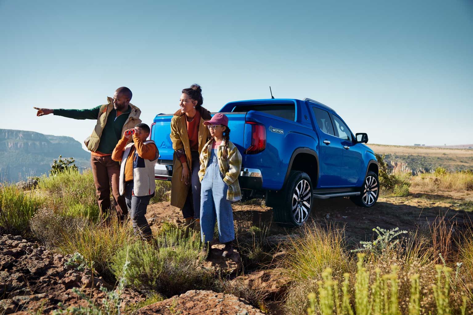 Eine Familie parkt mit einem blauen Amarok in den Bergen und blickt in die Ferne