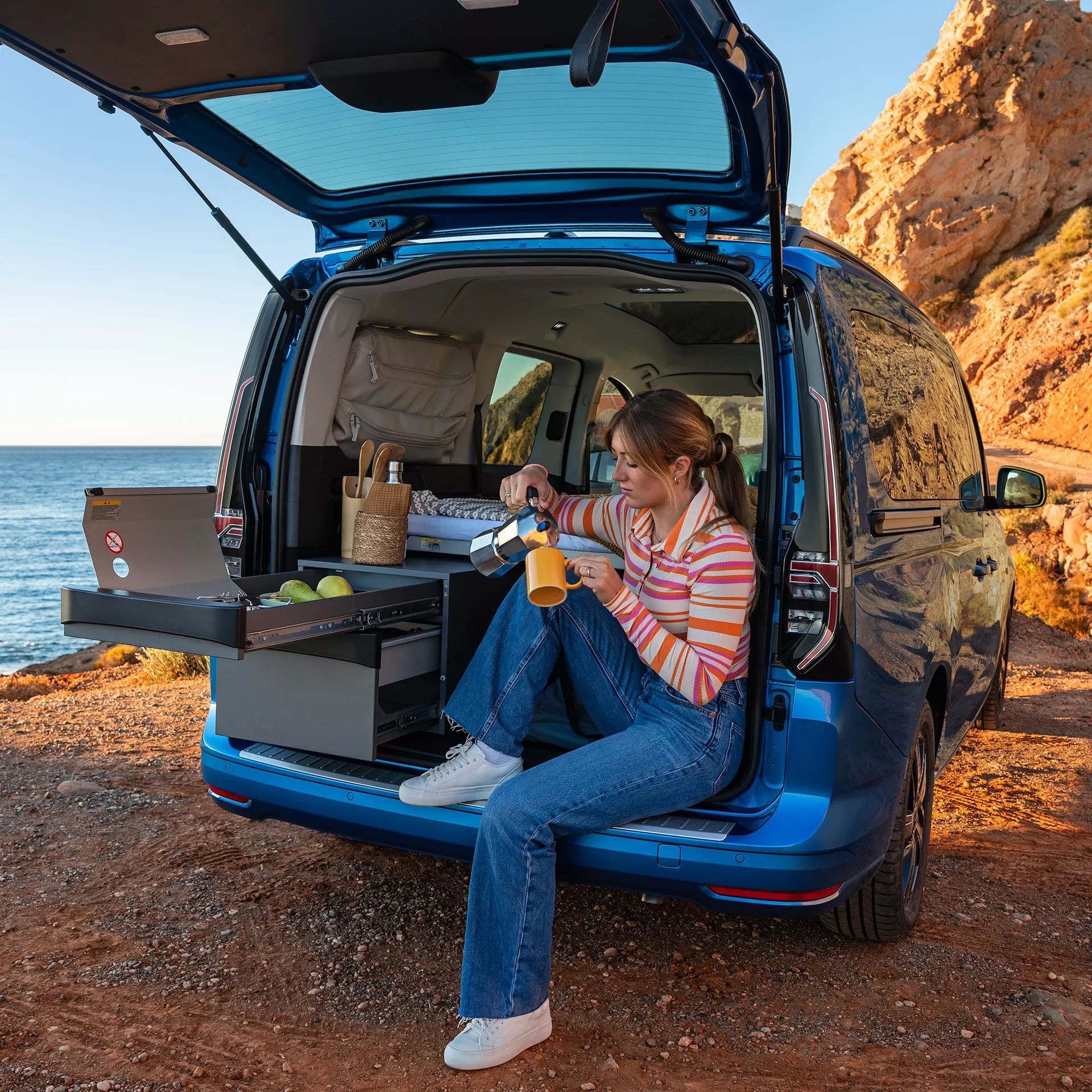 Eine Frau sitzt bei geöffneter Heckklappe in ihrem VW Caddy California, der am Meer parkt