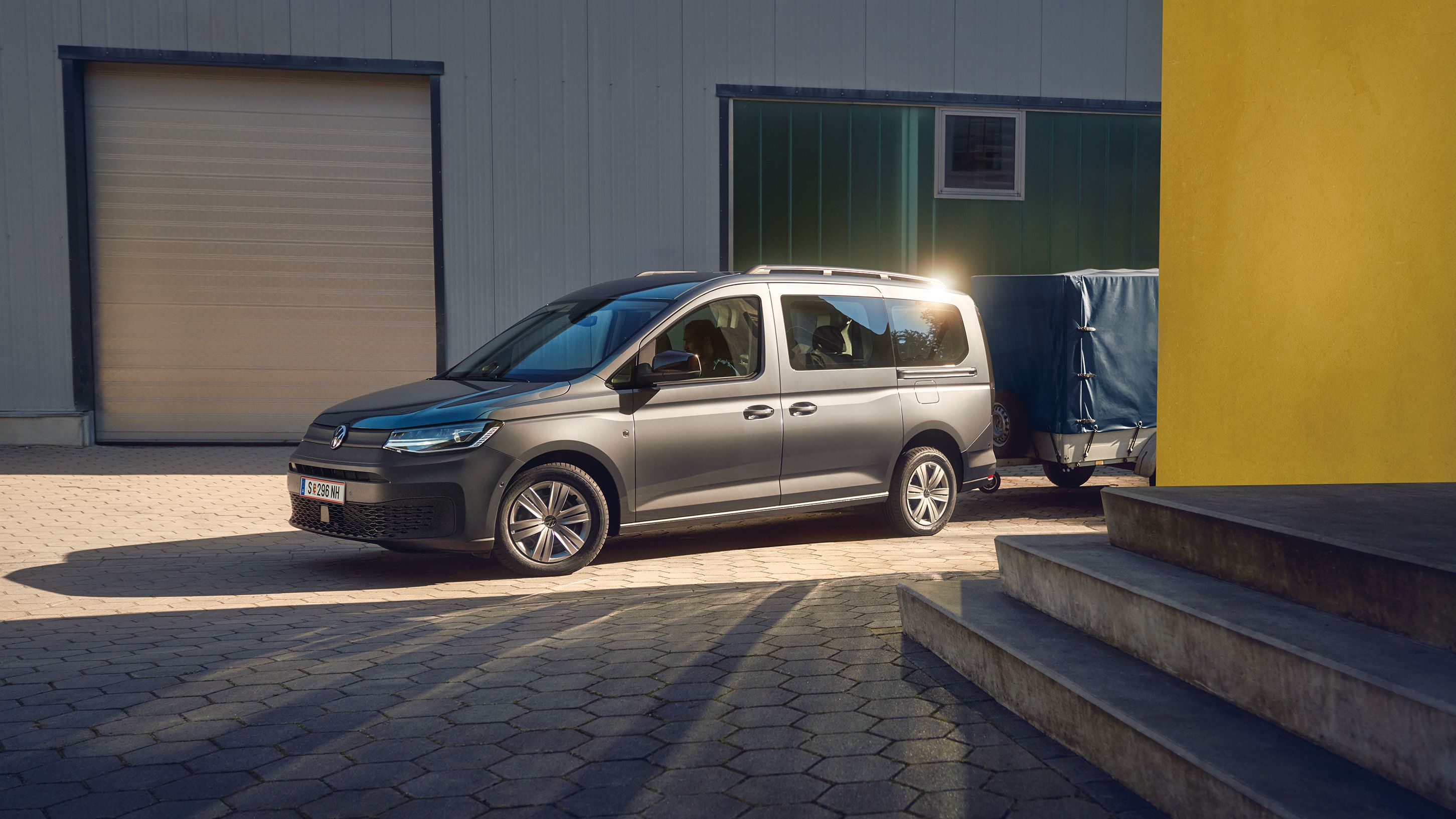 Ein grauer VW Caddy mit Anhänger parkt neben einer Garage