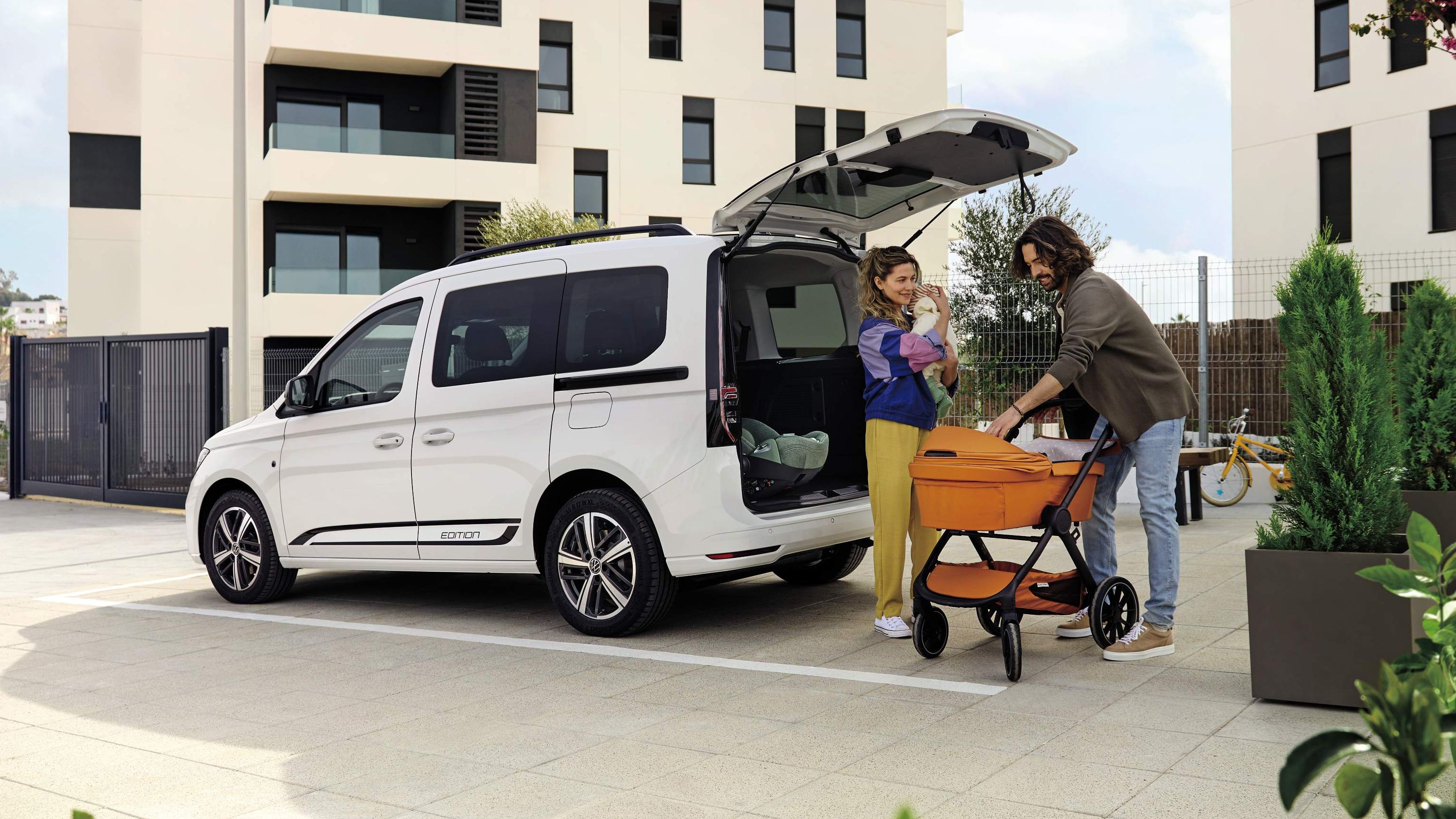 Eltern verstauen einen Kinderwagen im Kofferraum eines VW Caddy, Mutter hält das Baby