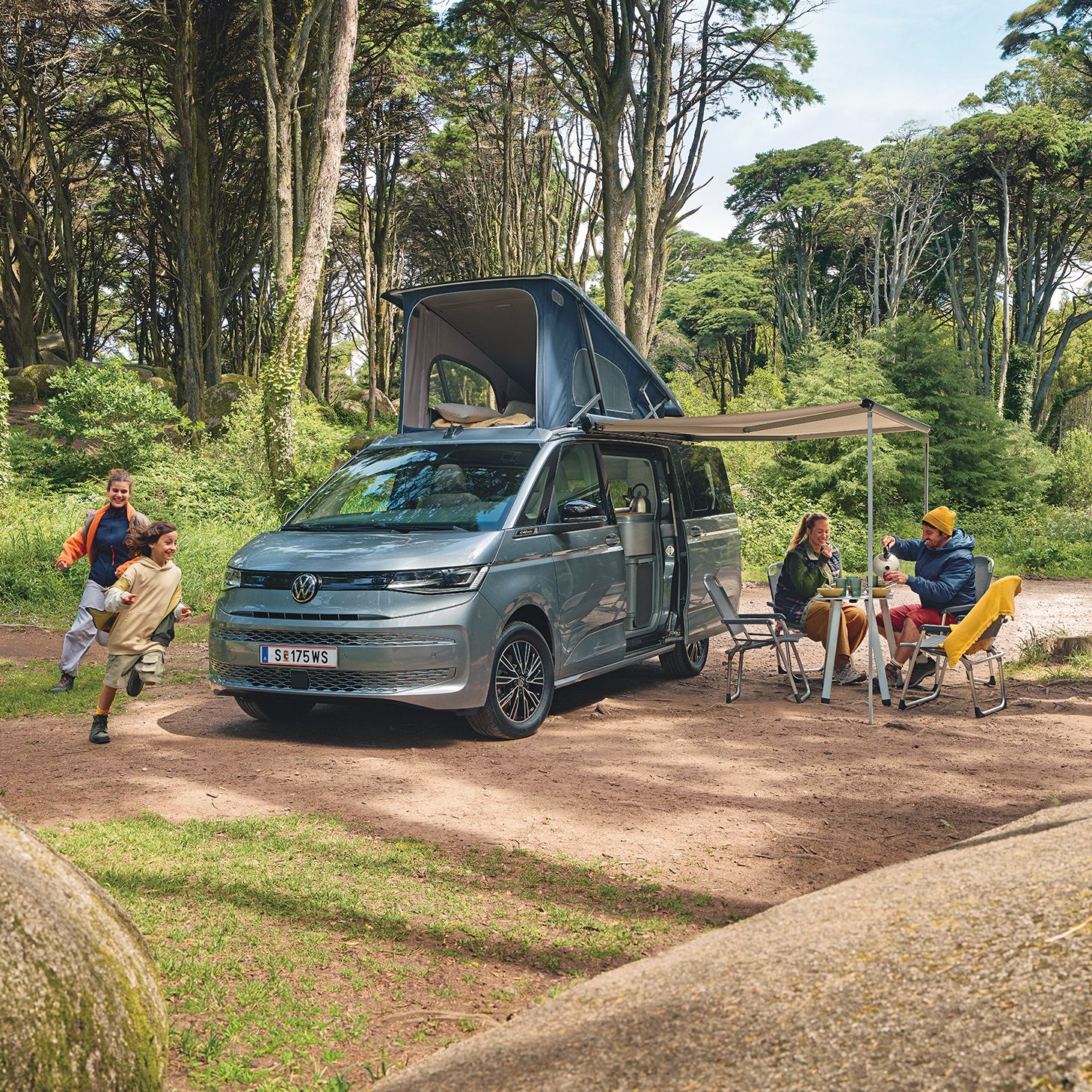 Ein New Multivan parkt im Wald,  zwei Kinder laufen darum herum, die Eltern frühstücken unter der ausgefahrenen Marquise